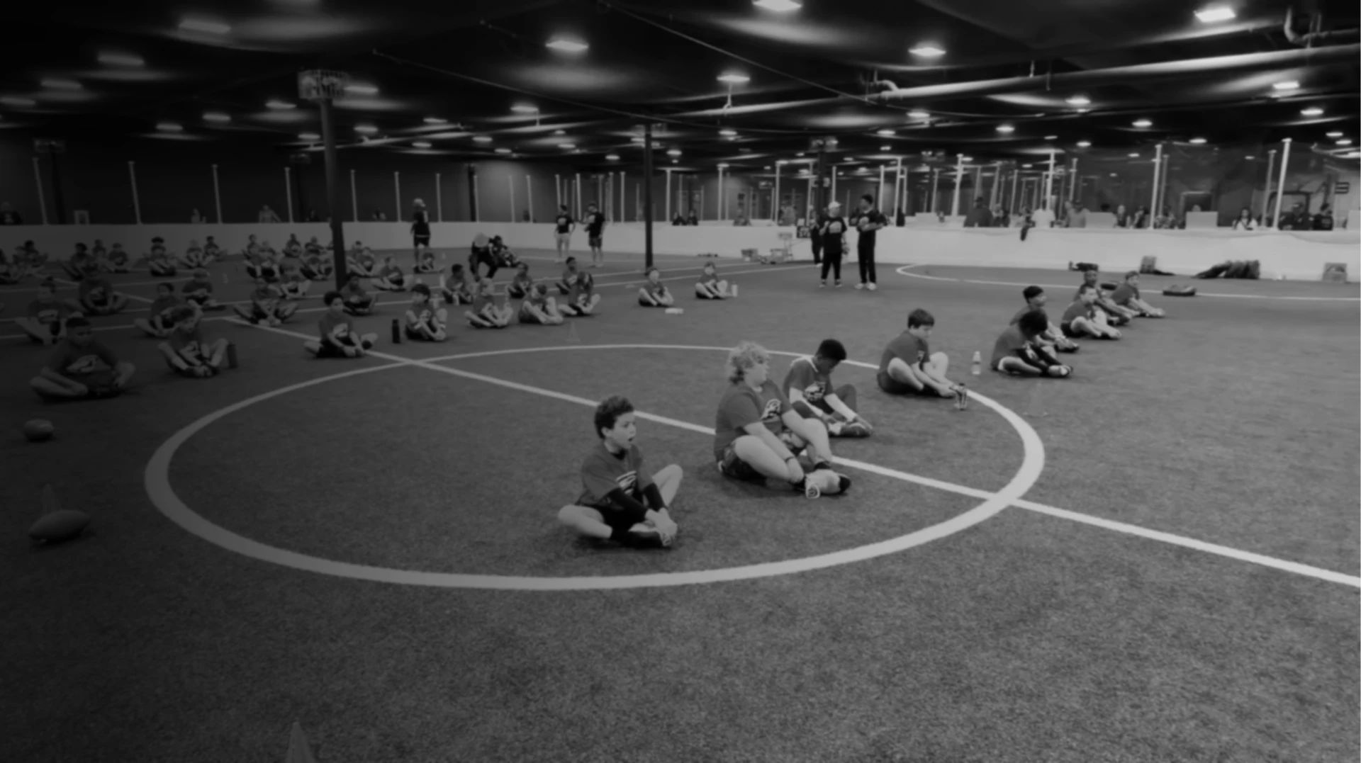 Campers seated on the indoor turf field listening to coaches at the Chris Cole Youth Football Camp